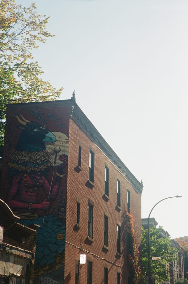 Brick apartment building with a large mural of a horned, bird-headed figure under a pale sky in Montreal, Canada