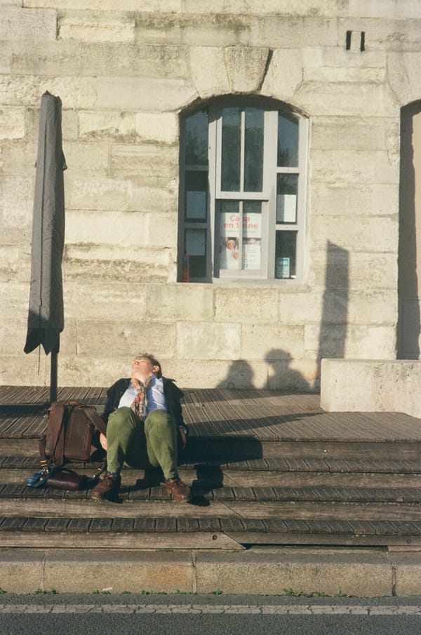 Man resting with eyes closed on sunlit wooden steps against a stone wall, his leather bag beside him, in Paris, France