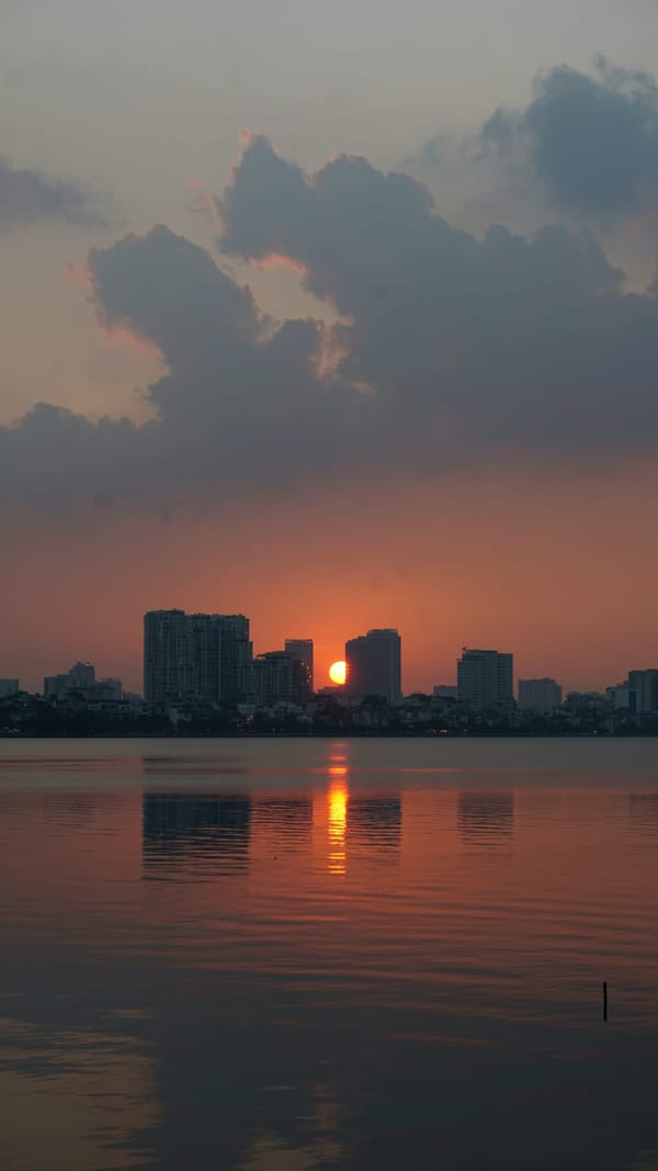 Orange sun setting between high-rise towers, its reflection glowing on a calm lake under heavy clouds in Hanoi, Vietnam