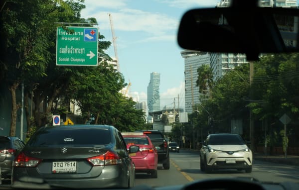 View through a car windshield of traffic on a tree-lined road below a green hospital sign in Bangkok, Thailand