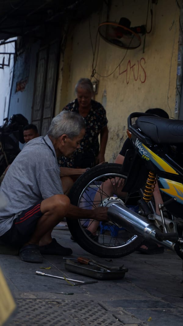 Older man crouching on the pavement repairing a motorbike's rear wheel as a woman looks on in Hanoi, Vietnam