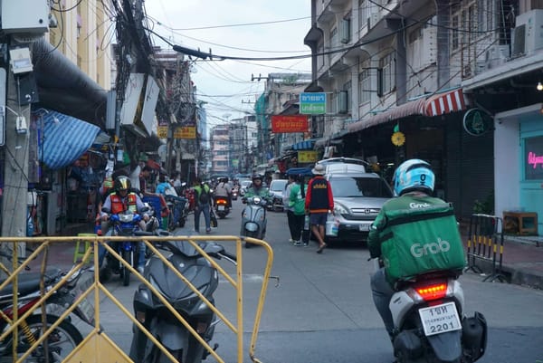 Grab rider and other motorbikes moving down a busy street of shophouses and tangled power lines in Bangkok, Thailand