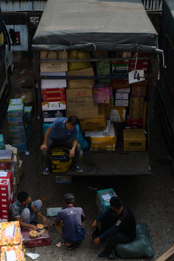 Workers unloading boxes of fruit from a covered truck while others sit and cut melons on the ground in Hanoi, Vietnam