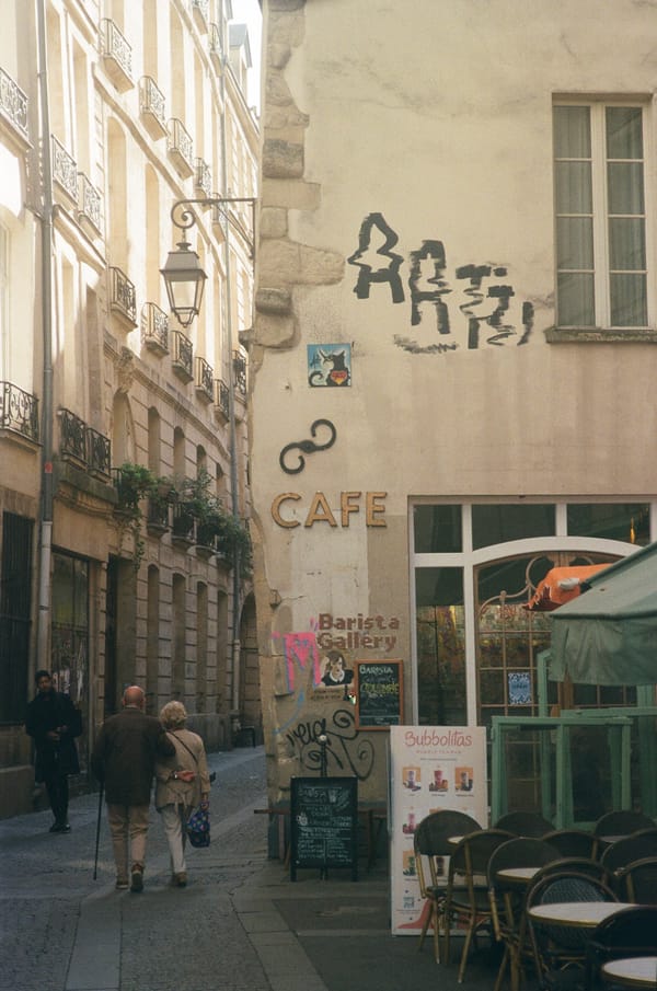 Elderly couple walking arm in arm down a narrow lane past a corner café with graffiti and wicker chairs in Paris, France