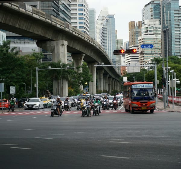 Motorbikes and an orange bus waiting at a red light beneath an elevated rail line and high-rises in Bangkok, Thailand