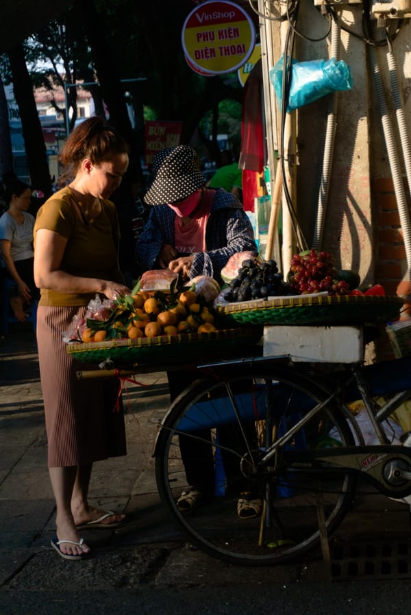 Woman choosing oranges from a fruit vendor's bicycle cart in low golden sidelight on a street in Hanoi, Vietnam