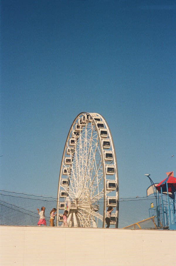 Children walking along a netted rooftop play area in front of a white Ferris wheel under clear blue sky in Montreal, Canada