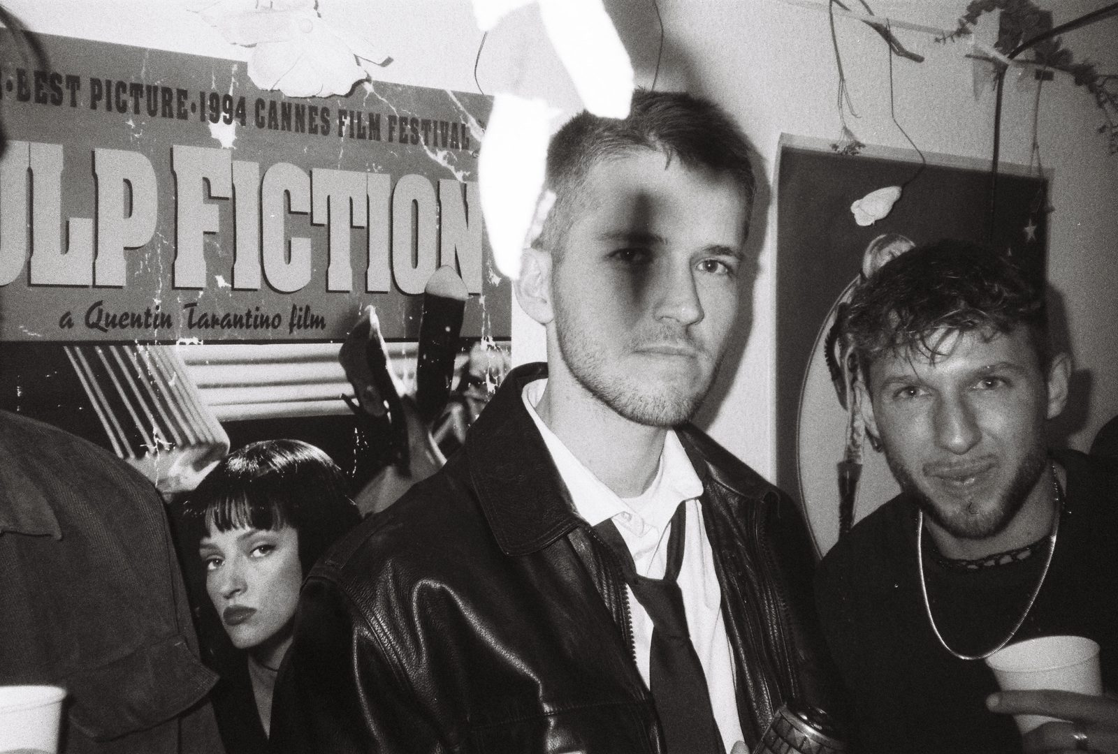 Black-and-white flash photo of partygoers in costume at a house party, a Pulp Fiction poster behind them