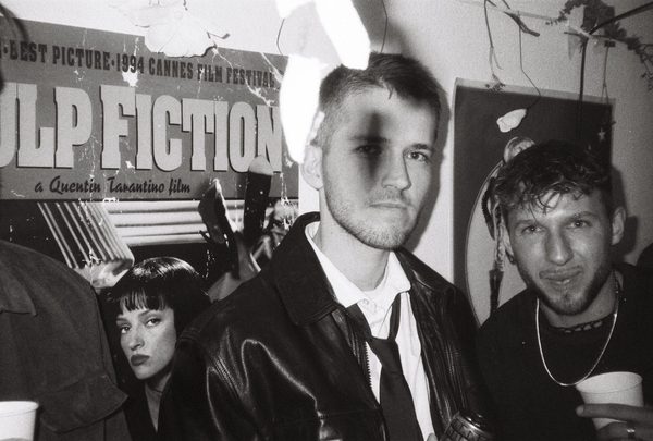 Black-and-white flash photo of partygoers in costume at a house party, a Pulp Fiction poster behind them