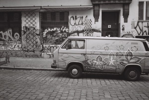 Black-and-white photo of a flower-painted camper van parked on cobblestones in front of graffiti-covered walls