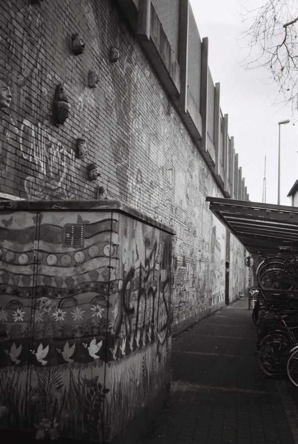 Black-and-white photo of a sidewalk along a brick wall covered in murals and masks, bicycles parked under a shelter