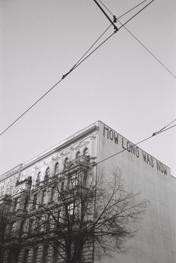 Black-and-white photo of tram wires crossing above an old facade painted with the words How Long Was Now