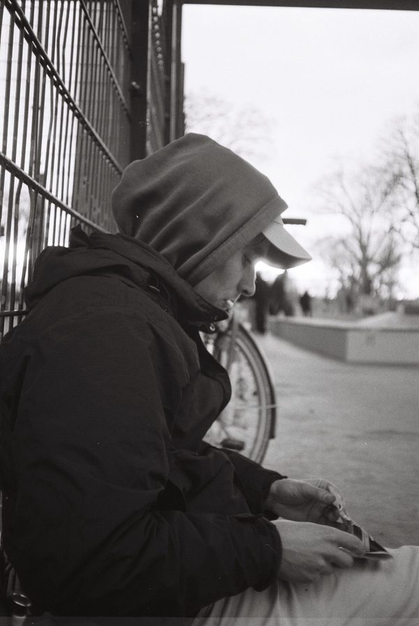 Black-and-white photo of a hooded young man in a cap sitting against a fence, looking down at something in his hands