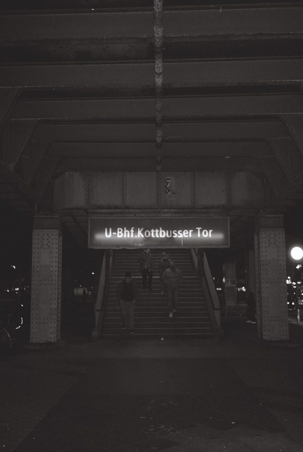 Black-and-white night photo of people descending the stairs beneath the U-Bhf. Kottbusser Tor sign in Berlin