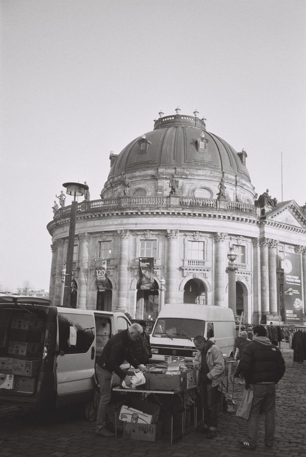 Black-and-white photo of market vendors unloading vans in front of the domed Bode Museum in Berlin