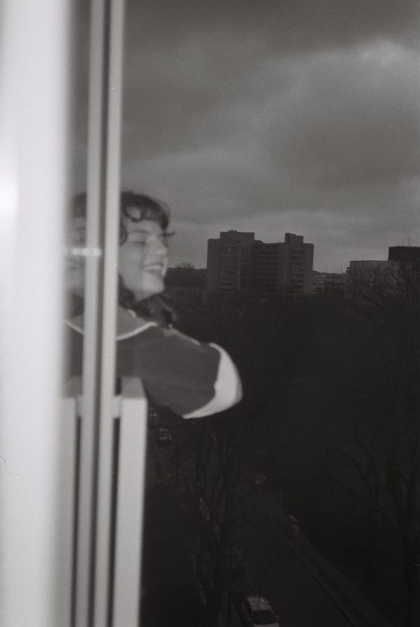 Black-and-white photo of a smiling young woman leaning out of a high window, apartment blocks under a heavy sky