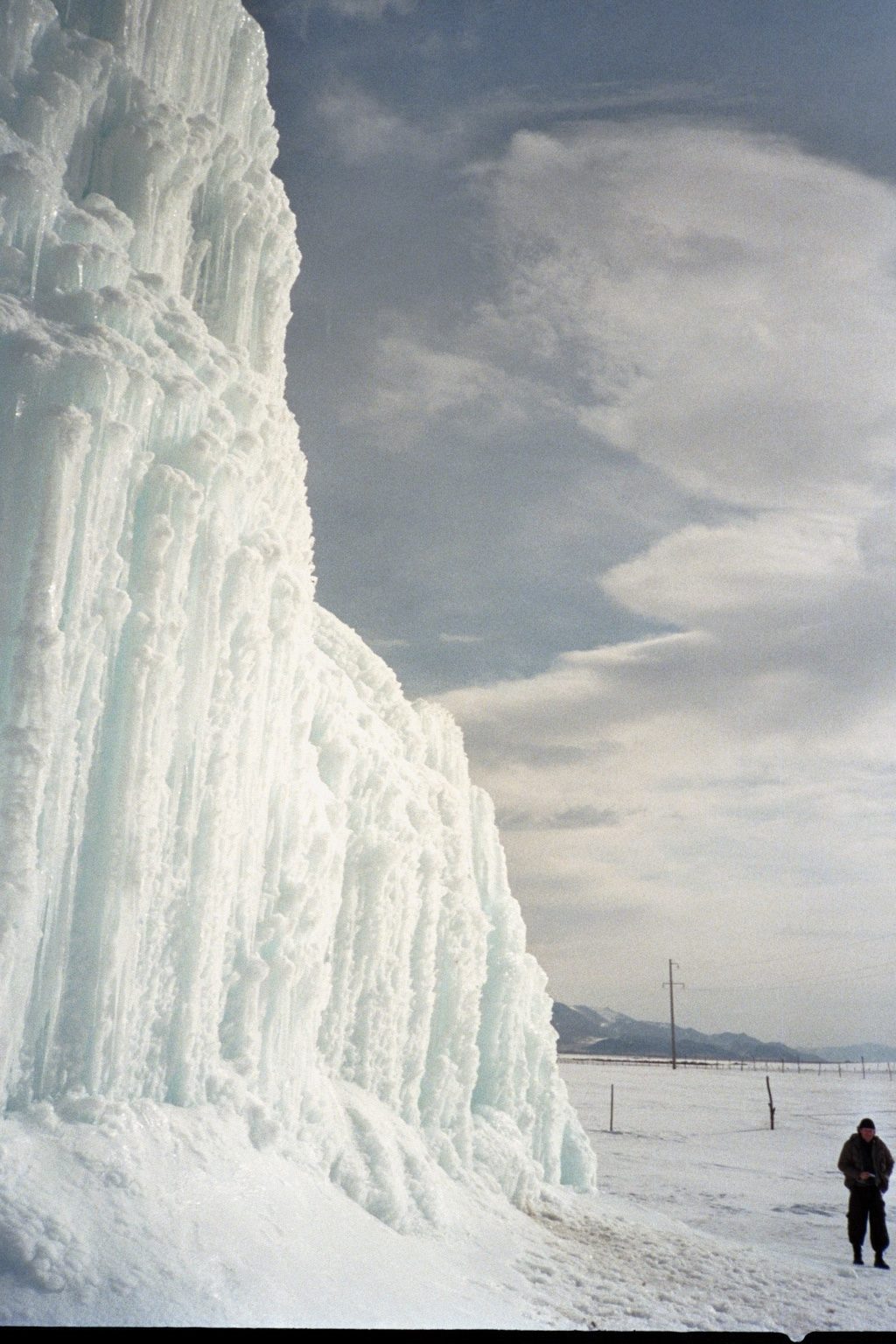 A towering wall of frozen icicles rises beside a snowy plain as a lone man stands in the cold, Central Asia