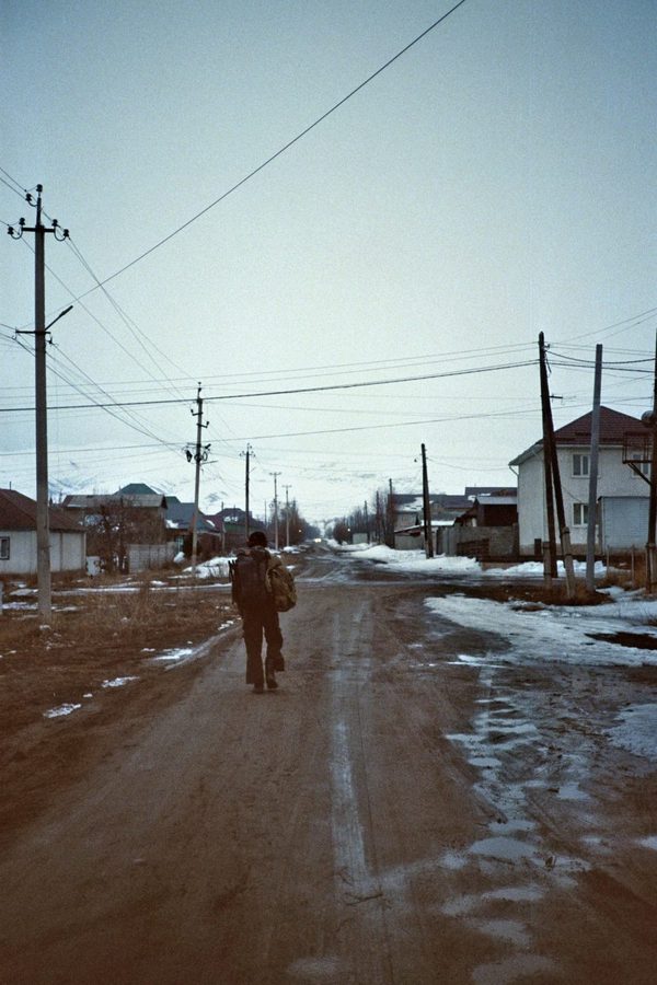 A backpacker walks down a muddy village road between houses and power lines with melting snow, Central Asia