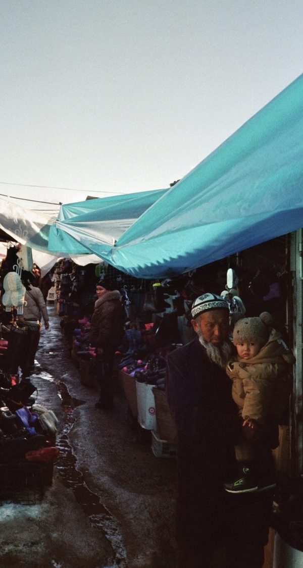 An older man with a white beard holds a toddler in a bear-ear hat at a market under blue tarps, Central Asia