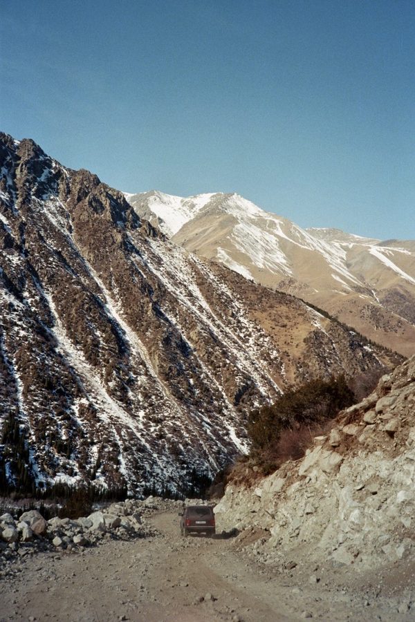 A small SUV drives along a rocky gravel track through a valley of snow-streaked mountains in Central Asia