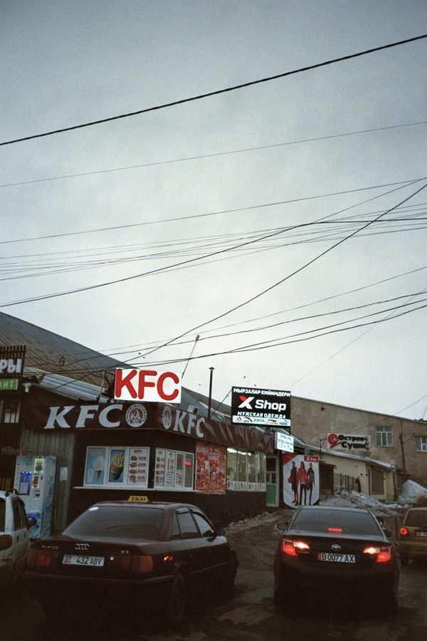 Cars wait outside roadside fast food and shop signs under a tangle of power lines on an overcast day, Central Asia