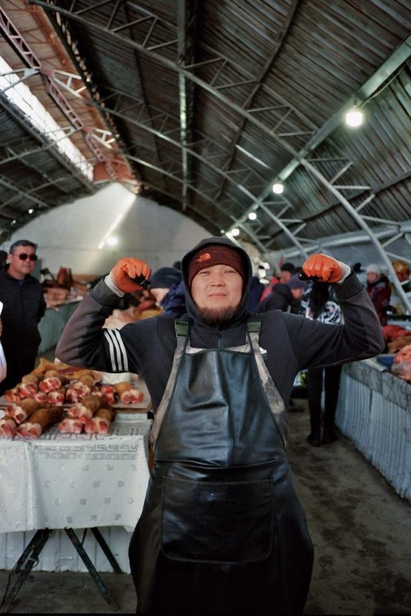 A smiling butcher in a leather apron flexes both arms in orange gloves inside a covered meat market, Central Asia