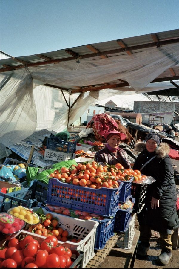 Two women in warm hats sell crates of tomatoes and tangerines under a plastic awning at a market in Central Asia