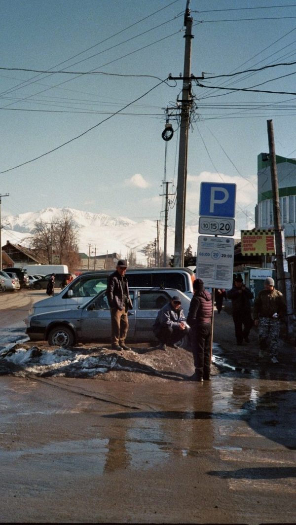 Men gather by parked cars and a parking sign on a slushy street with snowy mountains behind, Central Asia