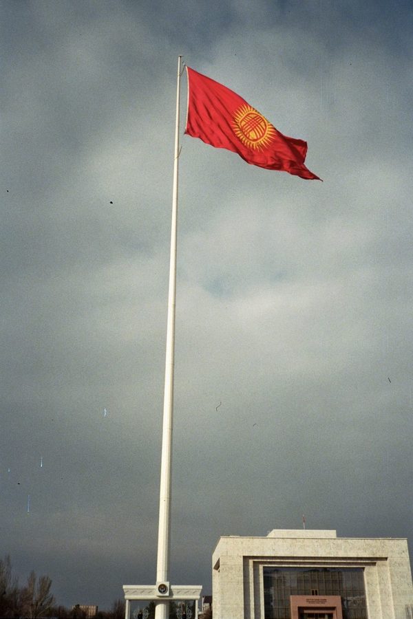 Kyrgyzstan's red flag with its yellow sun emblem flying from a tall white pole under a stormy grey sky, Central Asia