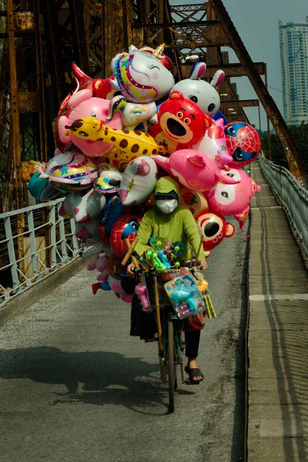 Masked vendor cycling across a rusty iron bridge under a huge cloud of cartoon foil balloons in Hanoi, Vietnam