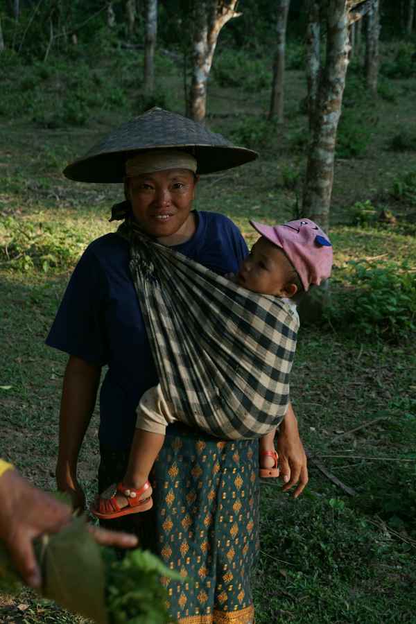 Woman in a conical hat carrying a toddler in a checked sling at the edge of a forest in Nong Khiaw, Laos