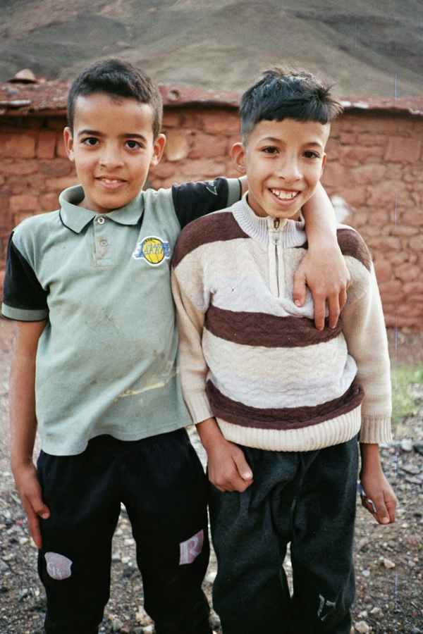 Two smiling boys standing arm in arm in front of a mud-brick wall and bare mountainside in Ighrem n'Ougdal, Morocco