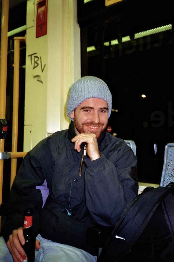 Smiling bearded man in a grey beanie sitting on a night tram with a bottle in his hand in Cologne, Germany