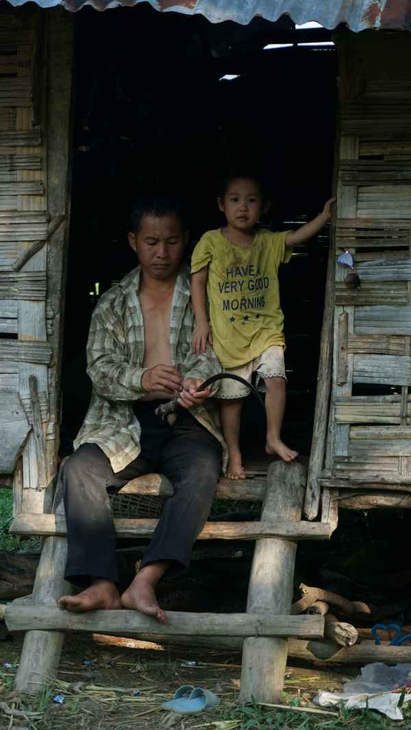 Man carving wood in the doorway of a bamboo house while a small child stands beside him in Nong Khiaw, Laos