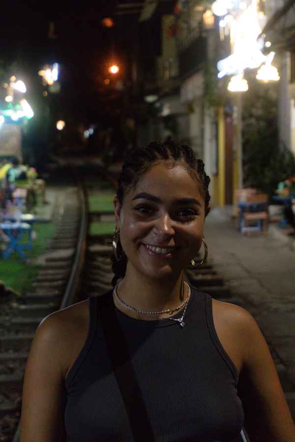 Smiling woman with braided hair and hoop earrings at night beside railway tracks lined with cafes in Hanoi, Vietnam