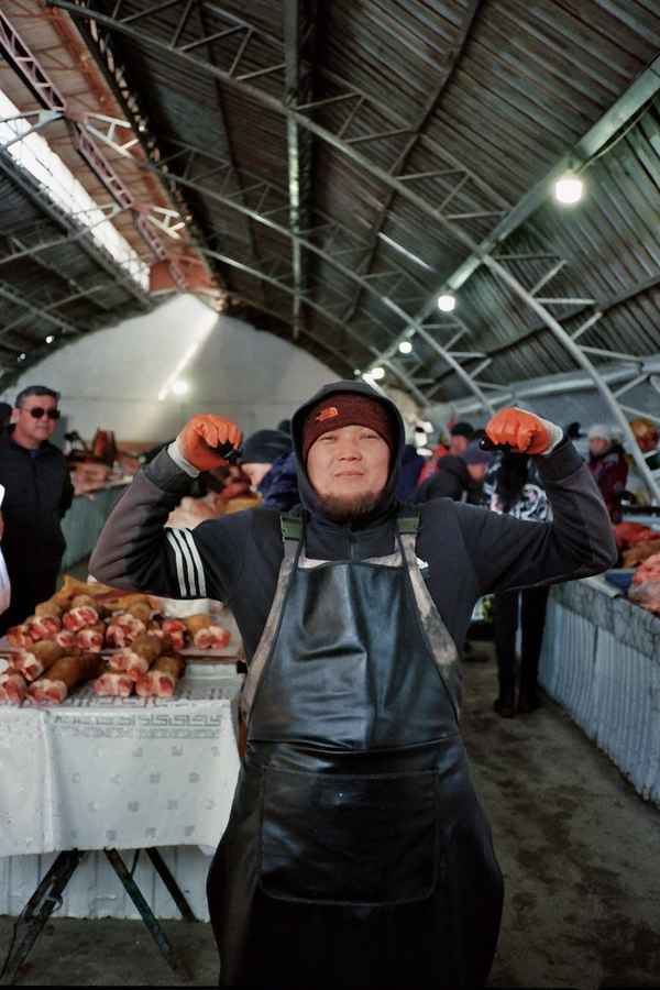 Market butcher in a leather apron flexing both arms in orange gloves inside a covered market hall in Karakol, Kyrgyzstan