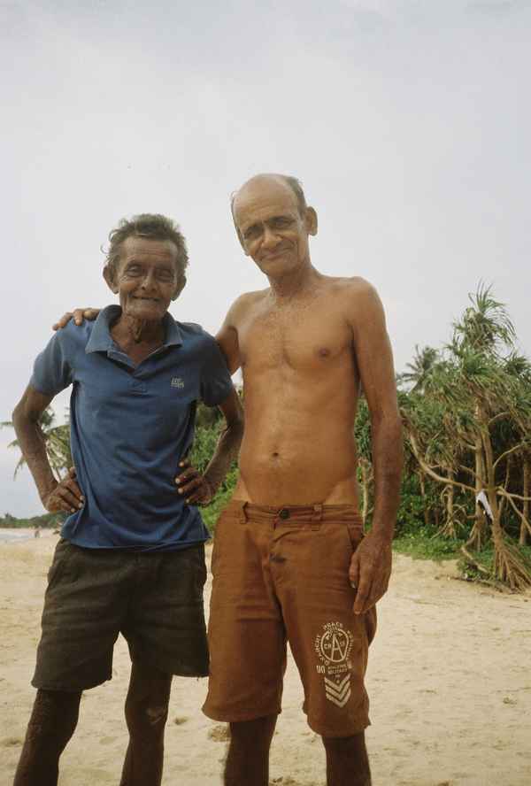 Two older men standing arm in arm on a sandy beach with pandanus trees behind them in Balapitiya, Sri Lanka