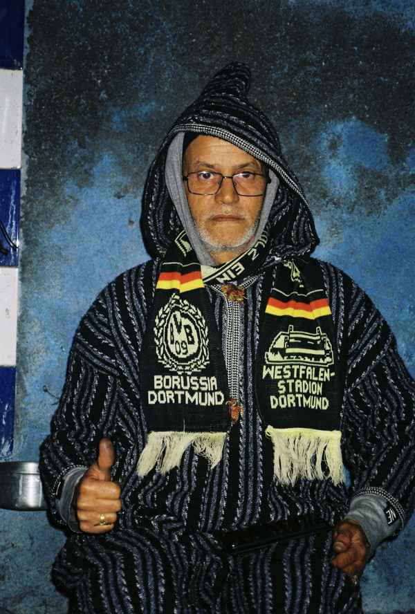 Man in a striped hooded djellaba and Borussia Dortmund scarf giving a thumbs up against a blue wall in Taghazout, Morocco