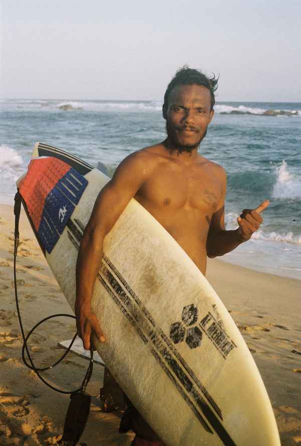 Shirtless surfer holding a worn surfboard on the beach in warm evening light, waves breaking behind, Midigama, Sri Lanka