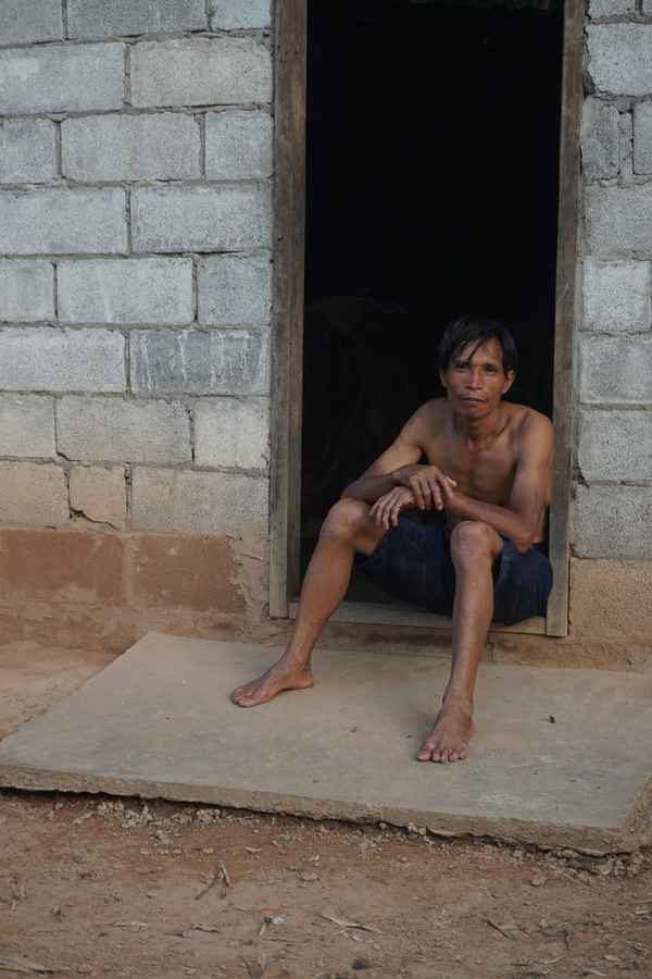 Barefoot man sitting in the doorway of a cinder-block house above a concrete step in Nong Khiaw, Laos
