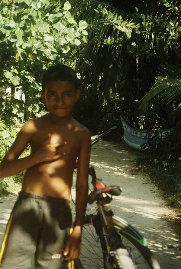 Shirtless boy with a bicycle on a sandy path under palms, an old blue boat in the shade behind, Balapitiya, Sri Lanka