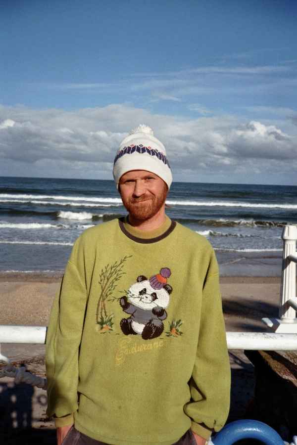 Bearded man in a bobble hat and panda sweater standing by a railing in front of the sea in Ribadesella, Spain