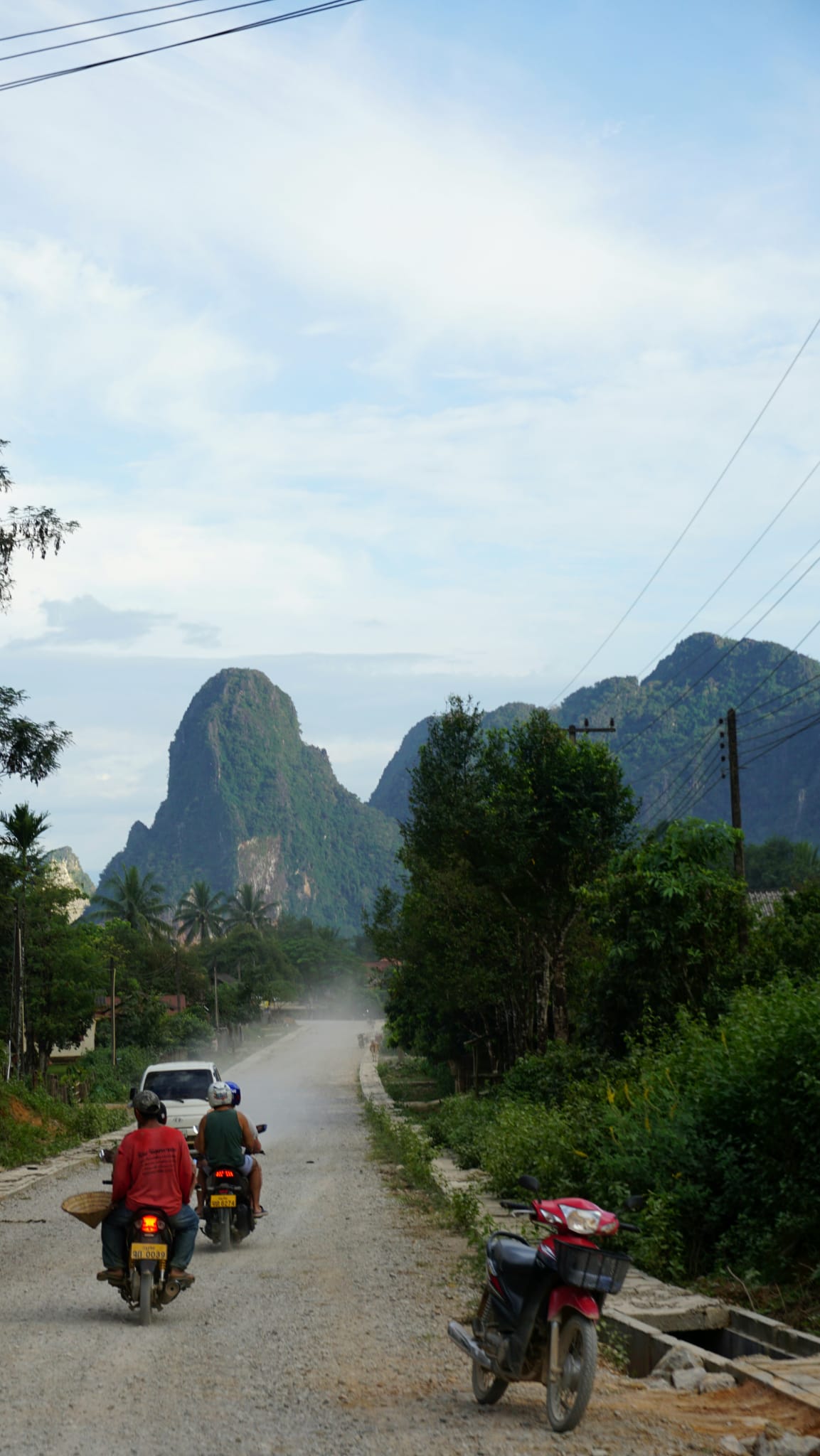 Motorbike riders on a dusty gravel road heading toward steep green karst mountains under a pale sky in Laos