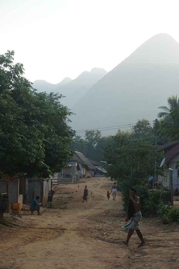 Villagers walking along a dusty dirt road between houses and trees, hazy mountain peaks rising behind, in Laos