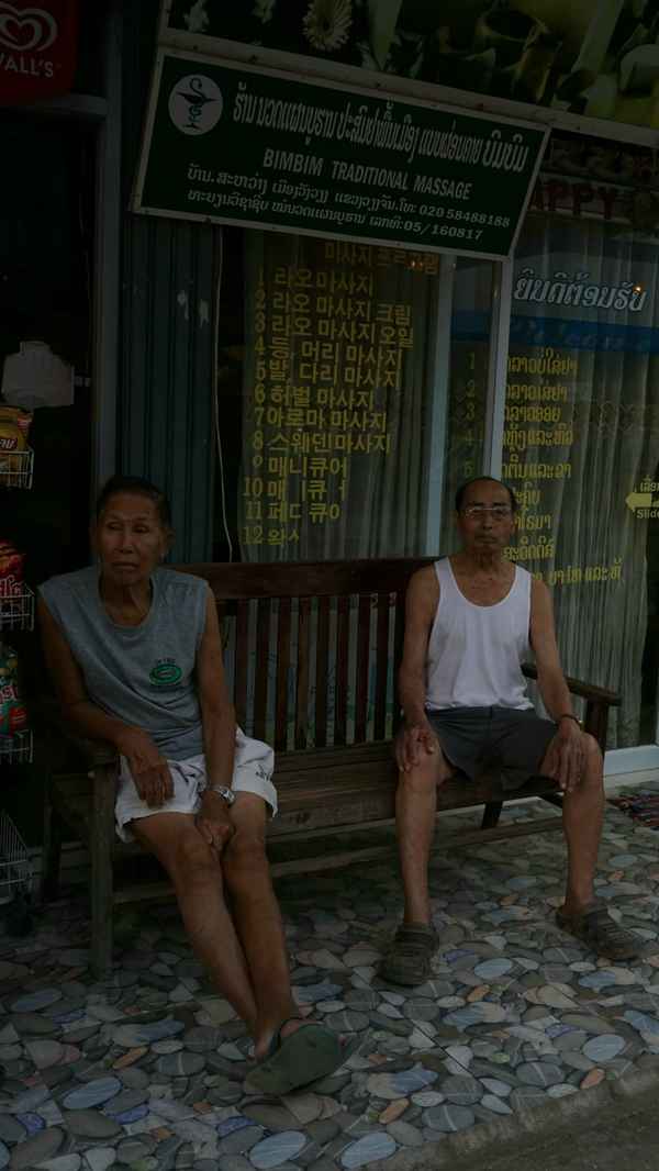 Two older men sitting on a wooden bench outside a massage shop with a multilingual price list in the window, Laos