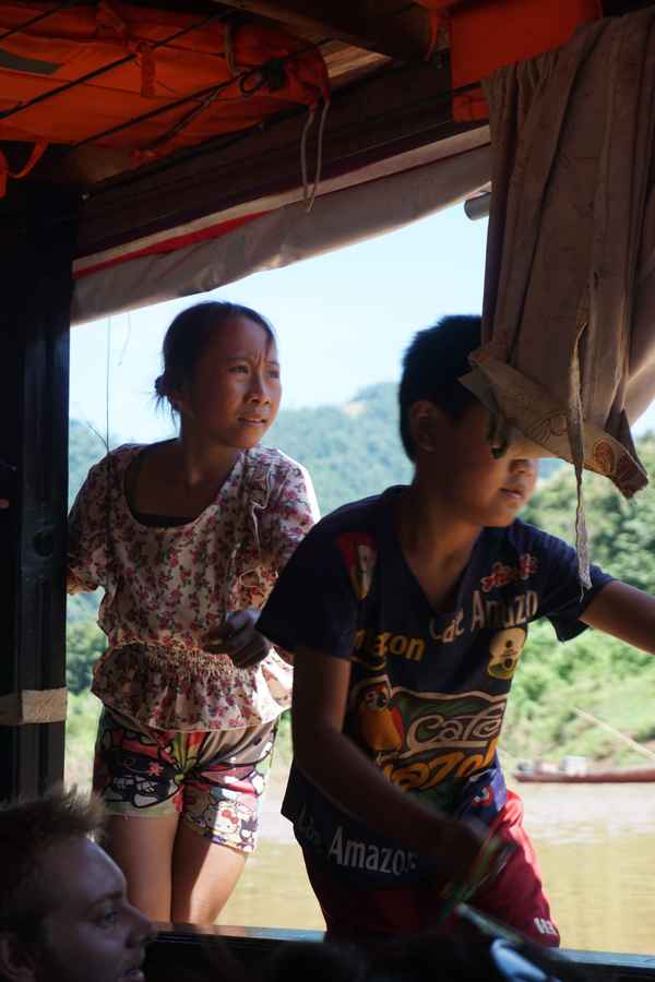 Girl and boy standing at the open side of a riverboat, looking out over brown water and forested hills in Laos