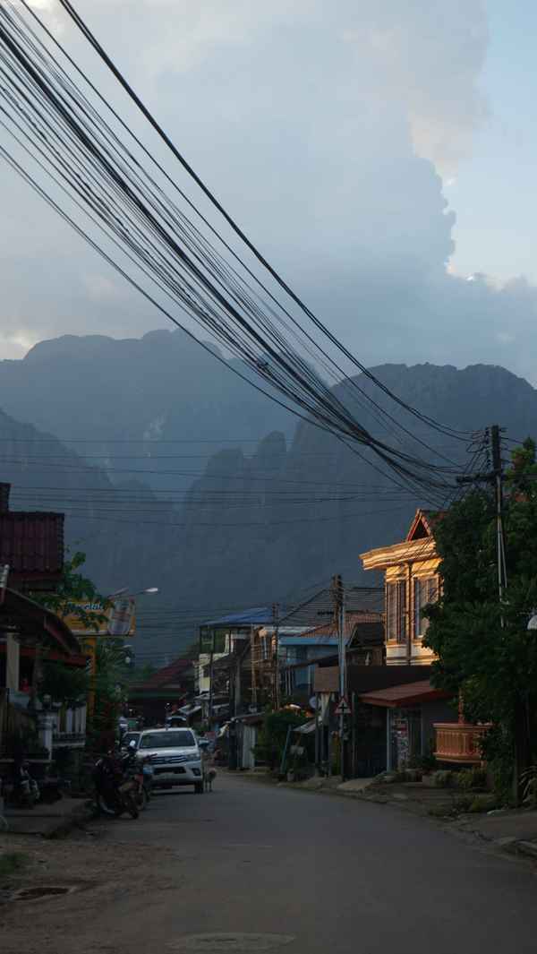 Quiet town street at dusk with dense overhead power cables and misty karst cliffs looming behind in Laos