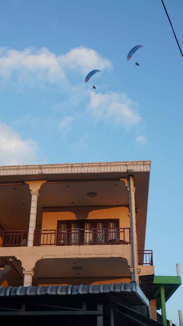 Two paramotor pilots flying in a blue sky with soft clouds above an orange two-storey house with balconies in Laos