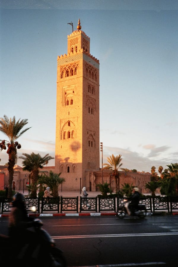 A tall stone minaret glows in warm evening light above palm trees as scooters blur past on the road, Morocco
