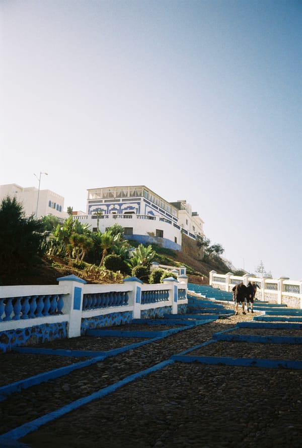 A blue and white staircase with painted railings climbs toward a white hilltop building under a clear sky, Morocco